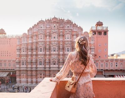 A woman in a floral dress stands on a balcony, looking at the intricate pink facade of Hawa Mahal palace under a bright sky.