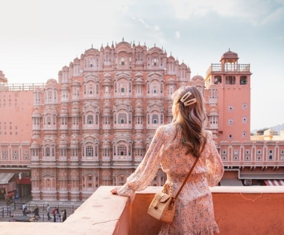 A woman in a floral dress stands on a balcony, looking at the intricate pink facade of Hawa Mahal palace under a bright sky.