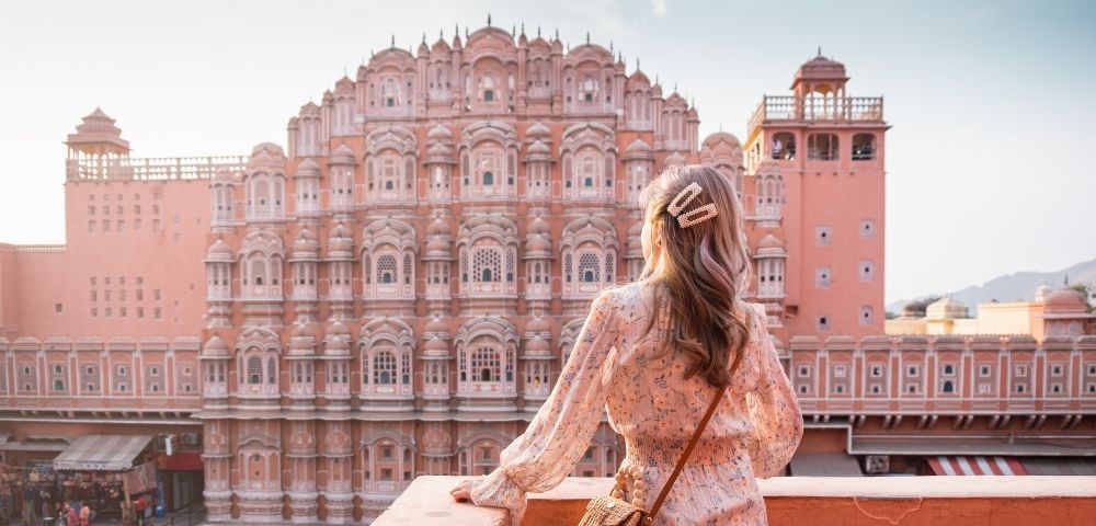 A woman in a floral dress stands on a balcony, looking at the intricate pink facade of Hawa Mahal palace under a bright sky.
