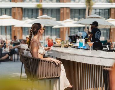 A person sits at a bar on a sunny day, holding a red cocktail.