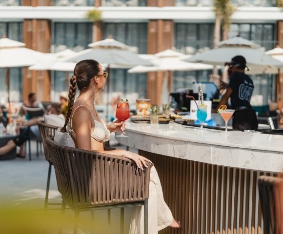 A person sits at a bar on a sunny day, holding a red cocktail.