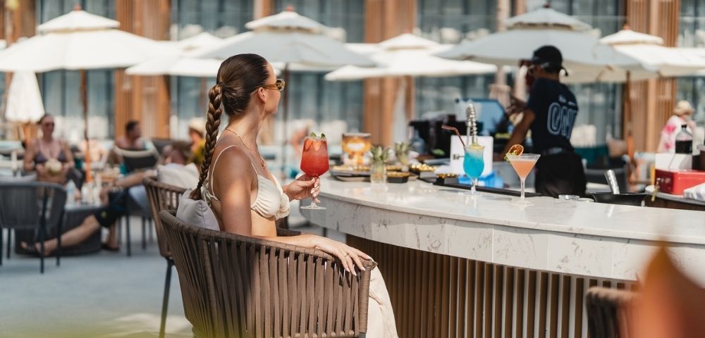 A person sits at a bar on a sunny day, holding a red cocktail.