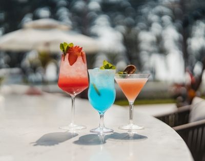 Three colorful cocktails sit on a marble bar in a tropical outdoor setting. The drinks are garnished with fruit and mint, and the background is softly blurred, showing umbrellas and seating.