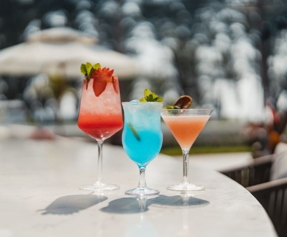 Three colorful cocktails sit on a marble bar in a tropical outdoor setting. The drinks are garnished with fruit and mint, and the background is softly blurred, showing umbrellas and seating.