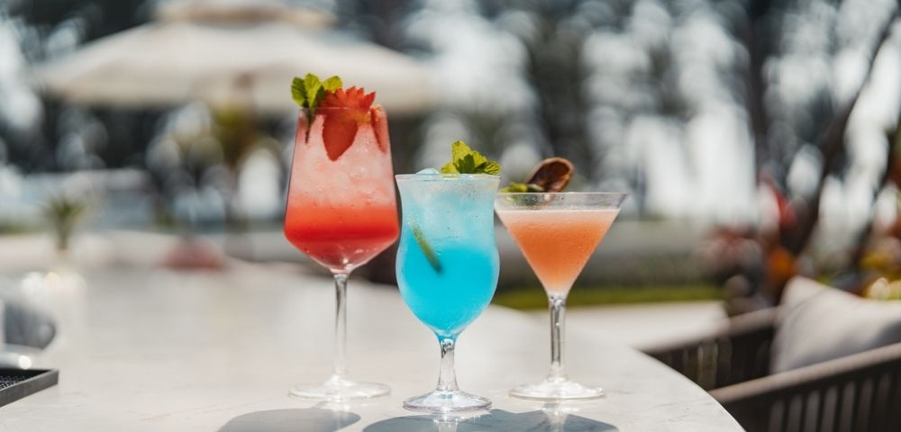 Three colorful cocktails sit on a marble bar in a tropical outdoor setting. The drinks are garnished with fruit and mint, and the background is softly blurred, showing umbrellas and seating.