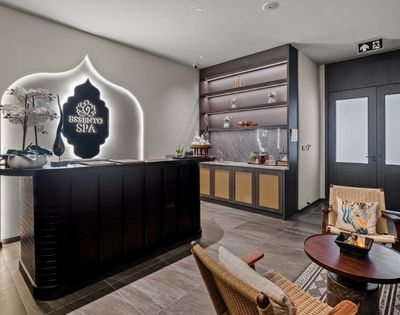 The reception area of a modern spa with a dark wood reception desk, a glowing spa logo, decorative orchids, and a counter with shelves displaying spa products.