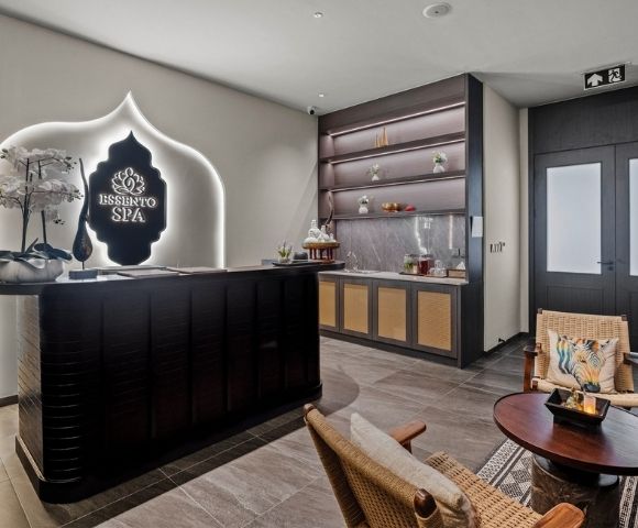 The reception area of a modern spa with a dark wood reception desk, a glowing spa logo, decorative orchids, and a counter with shelves displaying spa products.