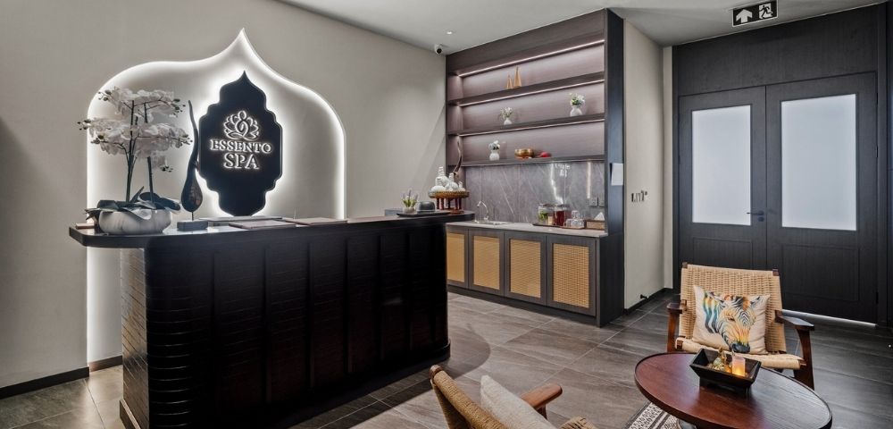 The reception area of a modern spa with a dark wood reception desk, a glowing spa logo, decorative orchids, and a counter with shelves displaying spa products.