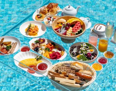 A lavish breakfast spread with various dishes like omelets, sausages, cheese platters, fruit bowls, pastries, and drinks, all served on a transparent table floating in a swimming pool.