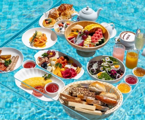 A lavish breakfast spread with various dishes like omelets, sausages, cheese platters, fruit bowls, pastries, and drinks, all served on a transparent table floating in a swimming pool.