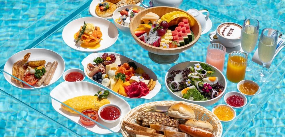 A lavish breakfast spread with various dishes like omelets, sausages, cheese platters, fruit bowls, pastries, and drinks, all served on a transparent table floating in a swimming pool.