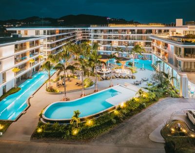 Luxury resort at night featuring illuminated swimming pools, palm trees, lounge areas, and modern multi-story buildings.