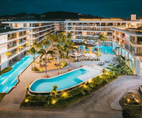 Luxury resort at night featuring illuminated swimming pools, palm trees, lounge areas, and modern multi-story buildings.