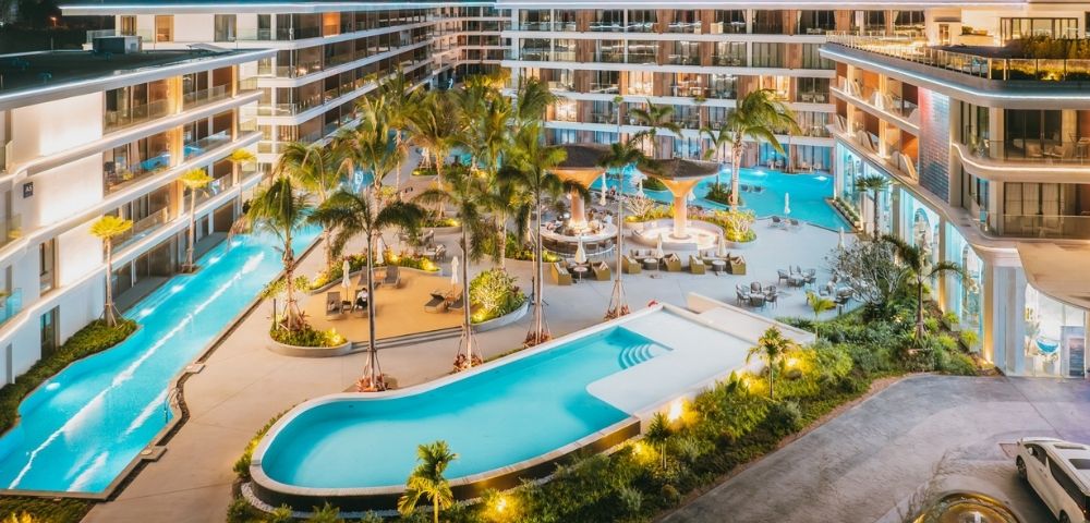 Luxury resort at night featuring illuminated swimming pools, palm trees, lounge areas, and modern multi-story buildings.