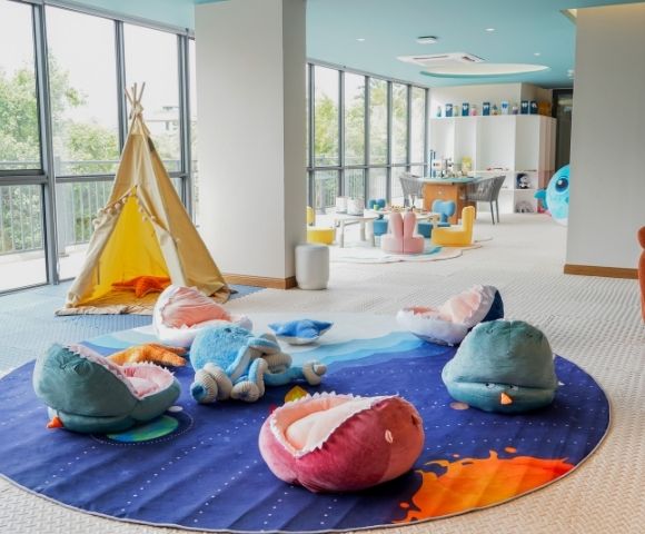 Bright children's playroom with a teepee tent, plush sea creature cushions, colorful chairs, and large windows letting in natural light.
