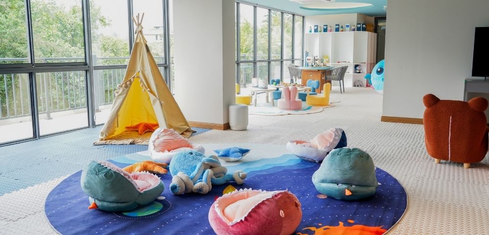 Bright children's playroom with a teepee tent, plush sea creature cushions, colorful chairs, and large windows letting in natural light.