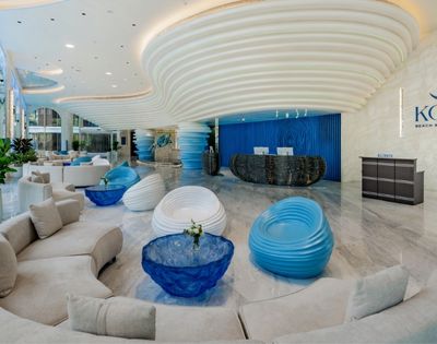 Modern luxury hotel lobby with curved beige sofas, blue and white sculptural chairs, glossy marble floor, and a wavy ceiling design at Kora Beach Resort.