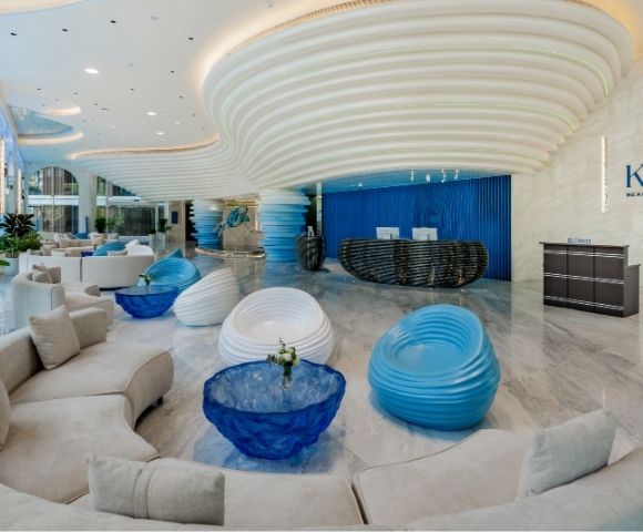 Modern luxury hotel lobby with curved beige sofas, blue and white sculptural chairs, glossy marble floor, and a wavy ceiling design at Kora Beach Resort.