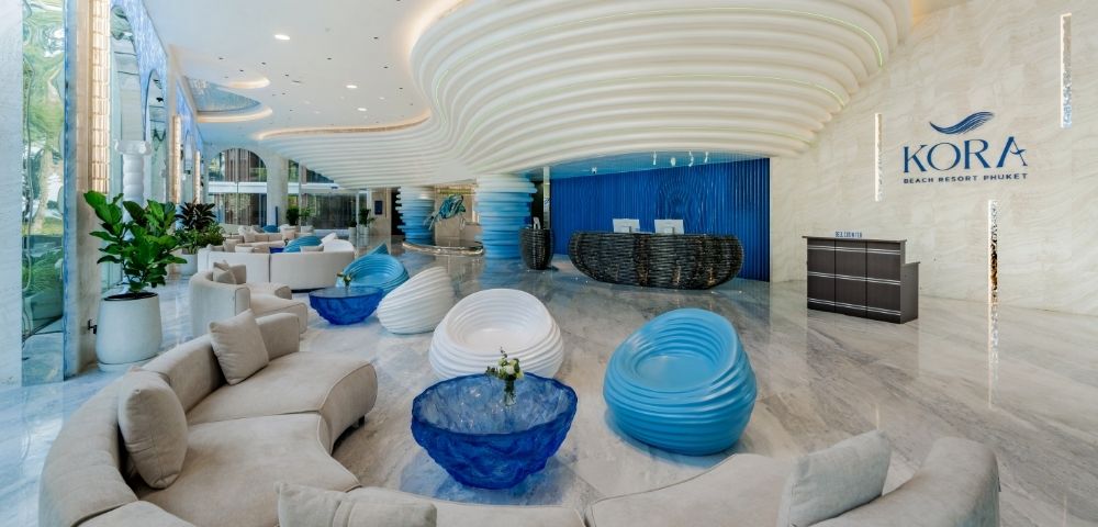 Modern luxury hotel lobby with curved beige sofas, blue and white sculptural chairs, glossy marble floor, and a wavy ceiling design at Kora Beach Resort.