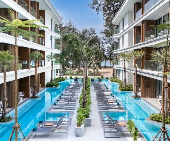 A modern hotel courtyard featuring a pool surrounded by lounge chairs and palm trees, with buildings on either side and a beach in the background.