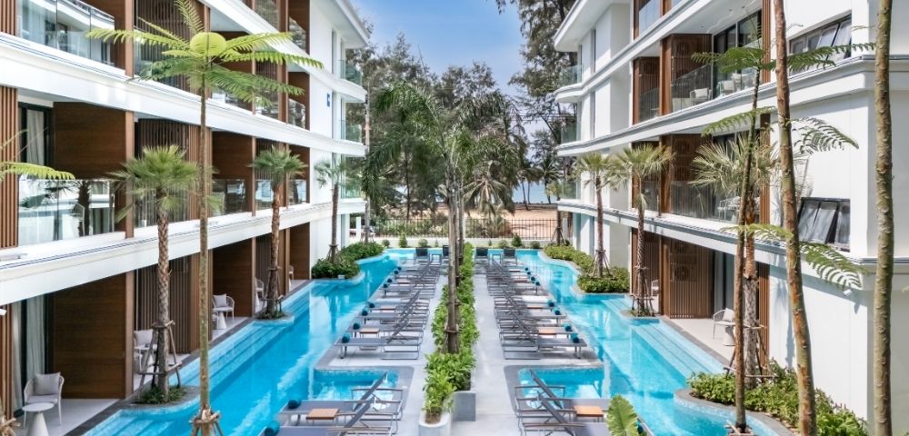 A modern hotel courtyard featuring a pool surrounded by lounge chairs and palm trees, with buildings on either side and a beach in the background.