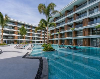 A modern hotel courtyard featuring a pool surrounded by lounge chairs and palm trees, with buildings on either side and a beach in the background.