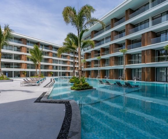 A modern hotel courtyard featuring a pool surrounded by lounge chairs and palm trees, with buildings on either side and a beach in the background.