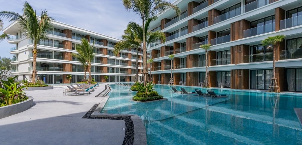 A modern hotel courtyard featuring a pool surrounded by lounge chairs and palm trees, with buildings on either side and a beach in the background.