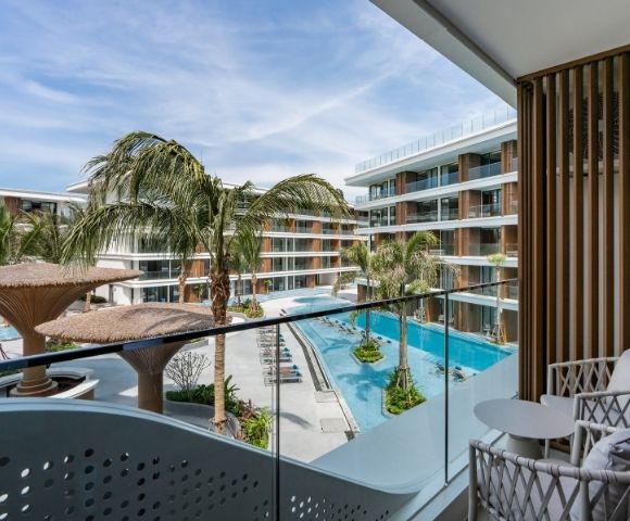 A view from a balcony overlooking a pool area surrounded by palm trees and modern hotel architecture.