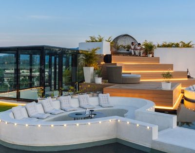 A luxurious rooftop terrace featuring modern seating, a sunken lounge area surrounded by water, and decorative lighting against a scenic backdrop.