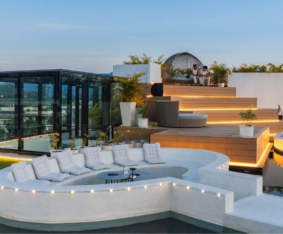 A luxurious rooftop terrace featuring modern seating, a sunken lounge area surrounded by water, and decorative lighting against a scenic backdrop.