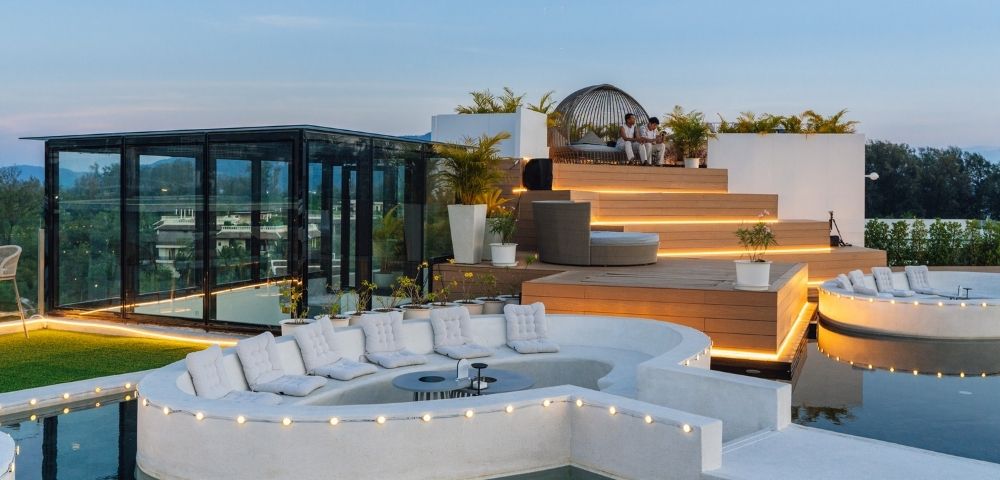 A luxurious rooftop terrace featuring modern seating, a sunken lounge area surrounded by water, and decorative lighting against a scenic backdrop.