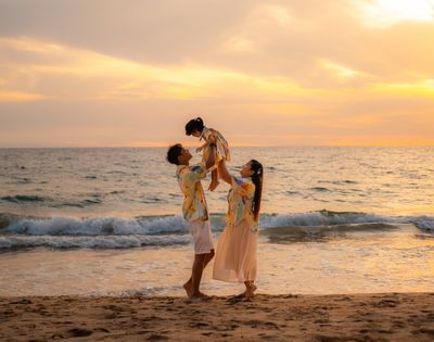 A family of three on the beach during sunset, with parents lifting their young child.