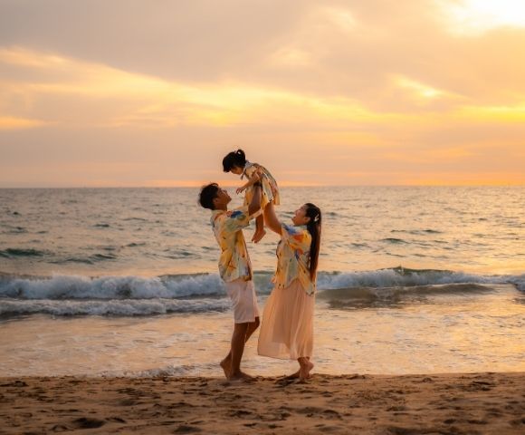 A family of three on the beach during sunset, with parents lifting their young child.