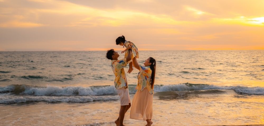 A family of three on the beach during sunset, with parents lifting their young child.