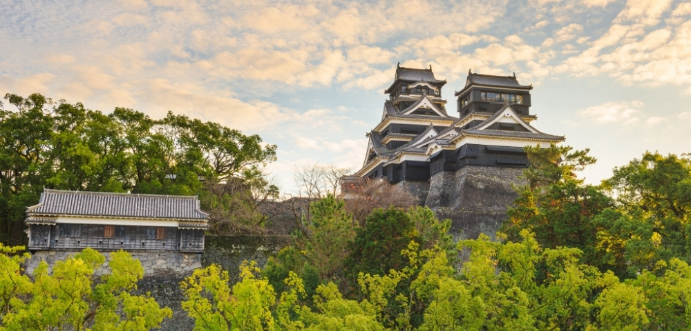 Historic Japanese castle with black-and-white tiered towers rises above stone walls and lush green trees beneath a softly clouded sky.