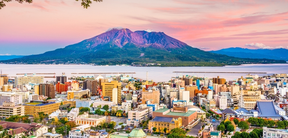 Panoramic cityscape beside a calm bay, with colorful buildings in the foreground and a dramatic volcanic mountain beneath a pink sunset sky.