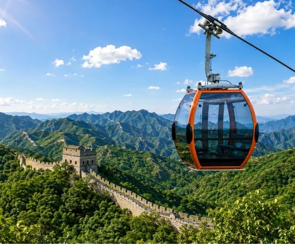 An orange cable car gondola is suspended in the foreground, with the Great Wall of China winding along a lush green, mountainous landscape under a bright blue sky with scattered clouds.