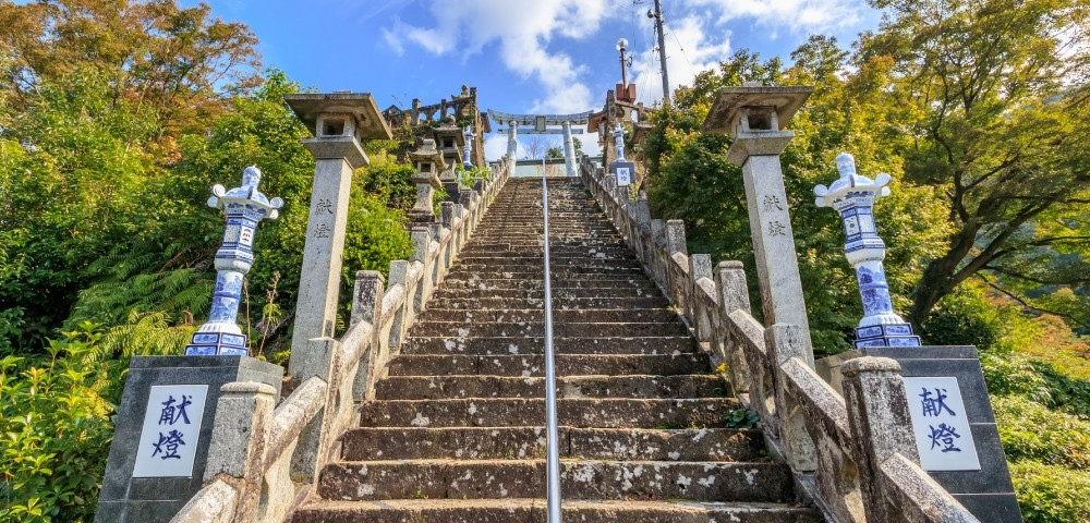 Steep stone staircase leading to a Japanese shrine, flanked by blue-and-white lanterns and carved pillars amid lush autumn trees.