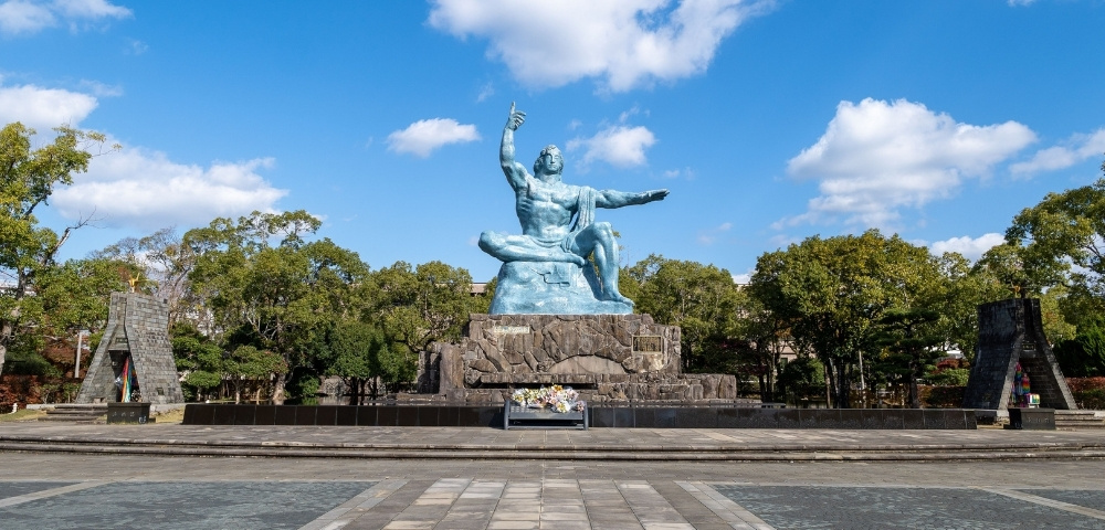 Monumental statue of a seated figure with outstretched arms, set on a stone pedestal in a tree-lined plaza beneath a bright sky.