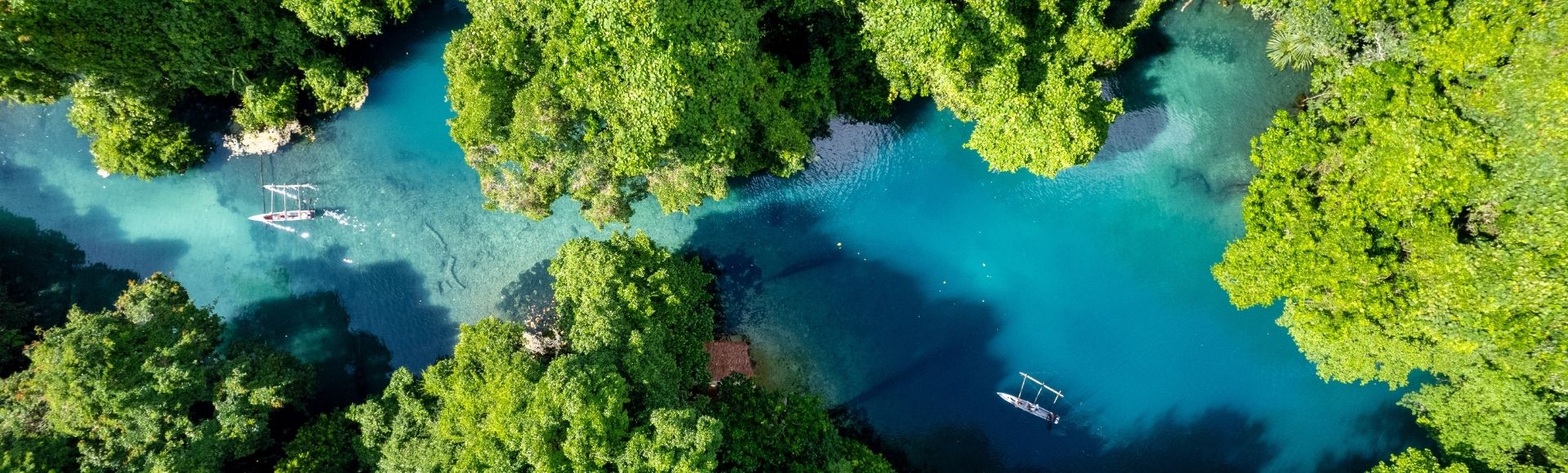 Aerial view of a vibrant blue river winding through a dense, lush green jungle, with two small outrigger canoes navigating the clear water.
