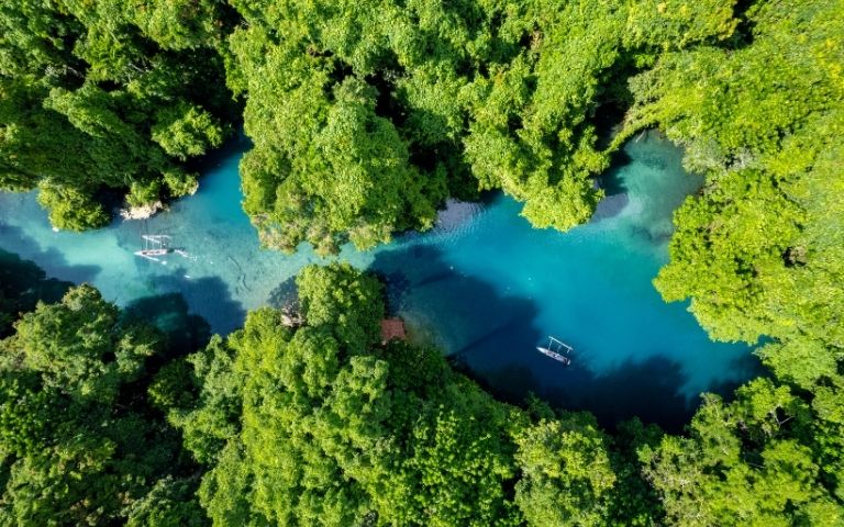 Aerial view of a vibrant blue river winding through a dense, lush green jungle, with two small outrigger canoes navigating the clear water.