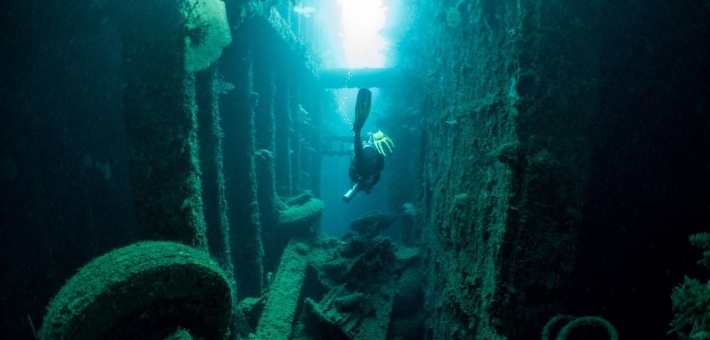 A solitary diver explores the interior of a sunken ship, illuminated by sunlight filtering through the water above, revealing the corroded metal walls and debris covered in marine growth.