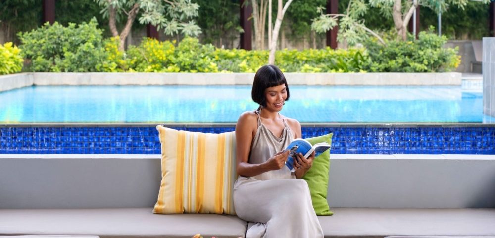 A woman sitting by a pool, reading a book, with a plate of pastries on the table beside her.
