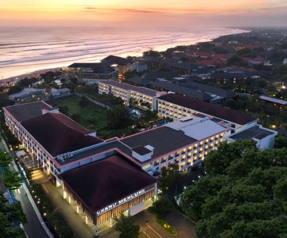 Aerial view of a hotel complex near a beach at sunset, with a mix of modern and traditional architecture surrounded by greenery and other buildings.
