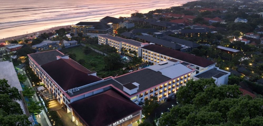 Aerial view of a hotel complex near a beach at sunset, with a mix of modern and traditional architecture surrounded by greenery and other buildings.