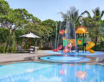 A water play area with colorful slides and a fountain, surrounded by green plants and a pool.