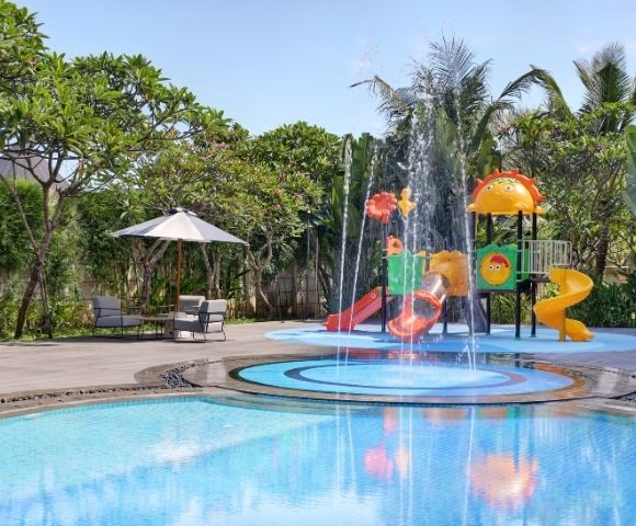 A water play area with colorful slides and a fountain, surrounded by green plants and a pool.