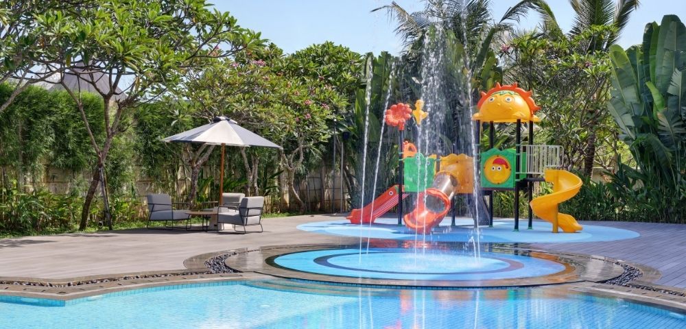 A water play area with colorful slides and a fountain, surrounded by green plants and a pool.