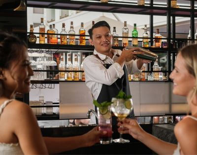 A bartender shakes a cocktail behind a bar while two women enjoy drinks and smile.